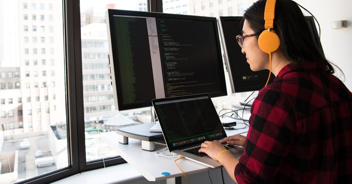 Woman programming on a laptop at a standing desk in an office with large windows.