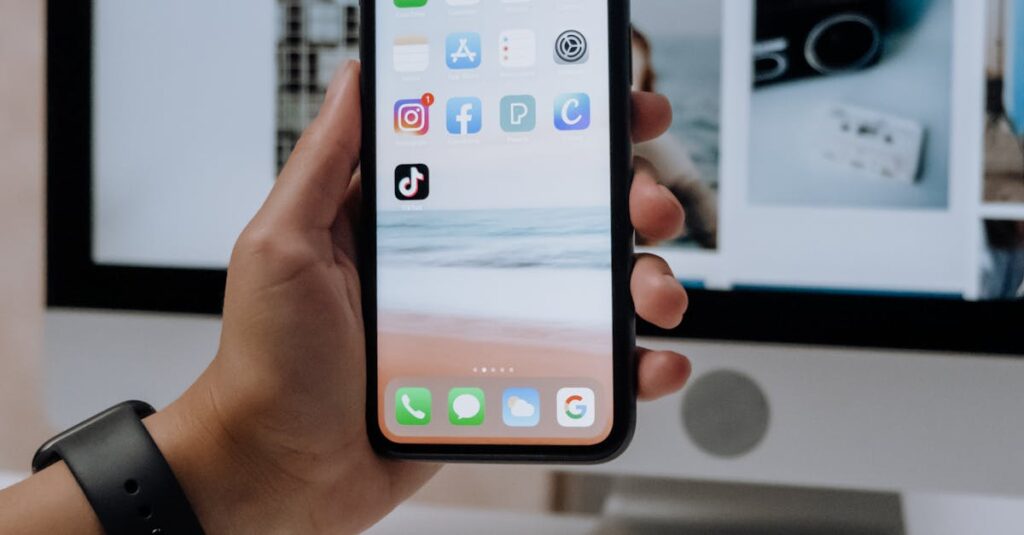 Close-up of a hand holding a smartphone showing social media apps in front of a desktop computer.